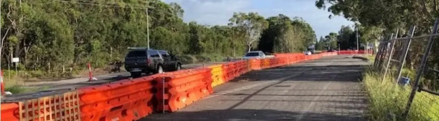 Orange construction barriers line a roadside, separating traffic from a closed lane with cars passing nearby.