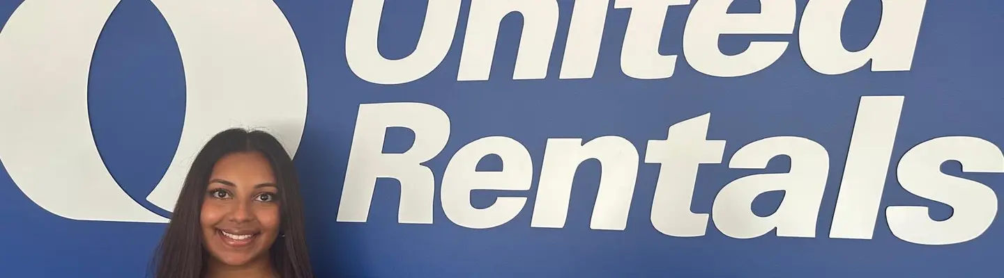 A woman standing in front of a blue wall with a big United Rentals logo on it.