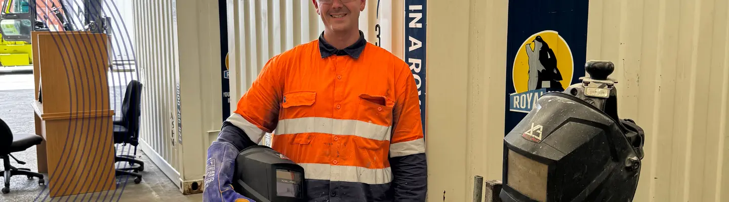 Person in hi-vis holding a welding helmet standing in front of a container.