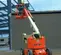 Rear view of an orange JLG articulating boom lift with extended boom and platform with a worker on a construction site.