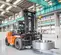 Front angled view of an orange and black Doosan warehouse forklift carrying a large coil attachment in a warehouse.