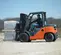 Side view of an orange warehouse forklift with a mast lifting pallets outdoors with an operator in the cabin.