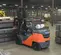 Rear angled view of an orange and grey Toyota warehouse forklift with an operator lifting pallets in a warehouse.