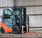 Front angled view of orange and grey Toyota warehouse forklift with a mast, orange forks and enclosed cabin in a warehouse.