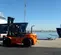 Side view of an orange and black warehouse forklift with a black mast positioned outdoors near water.