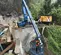 Side view of a blue boom lift with an extended arm lowering a worker on the platform over a ledge in a forested area.
