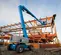 Rear angled view of blue articulating boom lift with extended arm working outdoors on orange steel scaffolding.