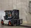 A white and black Bobcat D70 forklift with orange wheel hubs and black forks is driven by a man in an orange vest inside a warehouse with a concrete wall and floor. The forklift is shown from a front-left, slightly elevated angle.