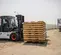 A white and black Bobcat D70 forklift with orange wheel hubs, lifting a large stack of wooden pallets on its black forks, is shown on a gravel lot with a white dome-shaped building and semi-trailers in the background under a clear sky. The forklift is viewed from its left side, slightly from the front.