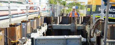 A construction worker in a hard hat and safety vest stands by a deep excavation, featuring heavy-duty metal shoring and rigging components, indicating complex installation work.