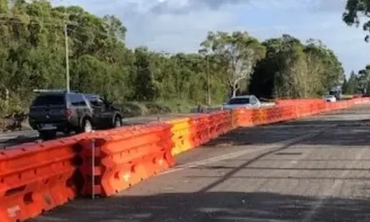 Orange construction barriers line a roadside, separating traffic from a closed lane with cars passing nearby.