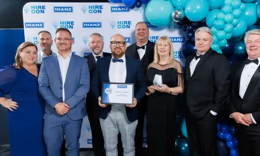 Employees smiling and holding awards, standing in front of a company logo backdrop at a recognition event.