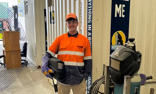 Person in hi-vis holding a welding helmet standing in front of a container.
